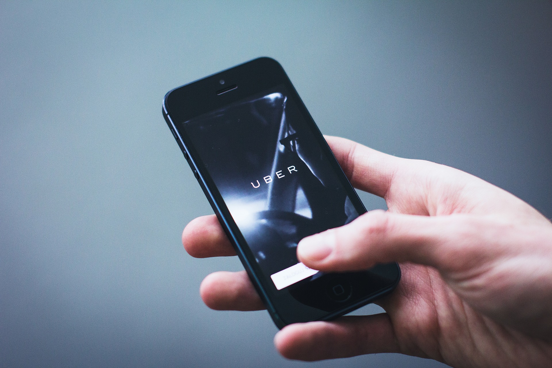 This is an image of a black cell phone being held by a hand coming in from the right-bottom of the image. The thumb is positioned over the screen about to touch a white tab at the bottom of the phone screen. On the screen is the word ‘UBER.' The background of the image is grey with shadowed corners.