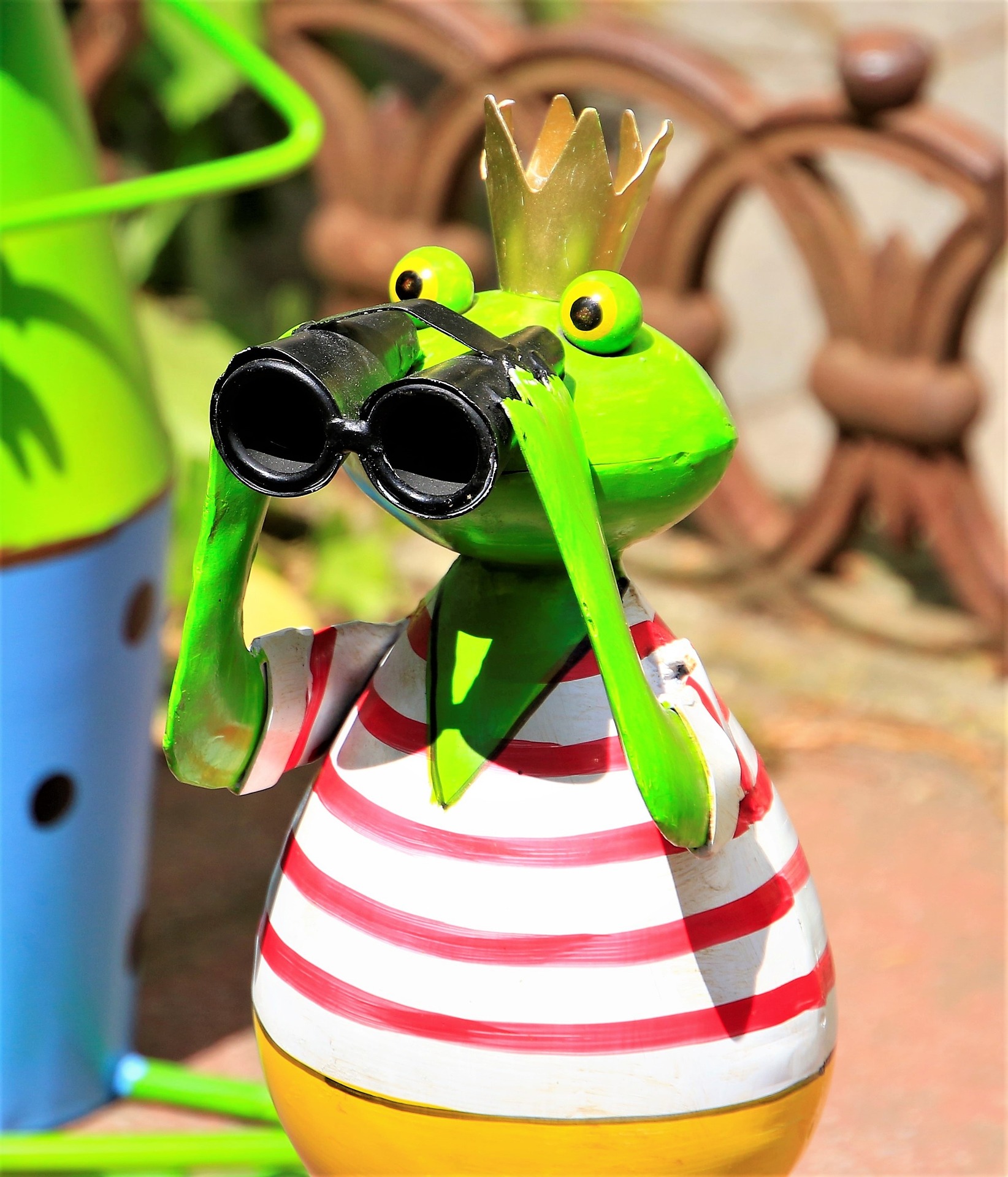This is an image of a green metal frog from the waist up. He is wearing yellow pants, a white shirt with horizontal red stripes, and a gold crown on his head. He is holding up black binoculars to his eyes. Other metal figures can be seen in the background.