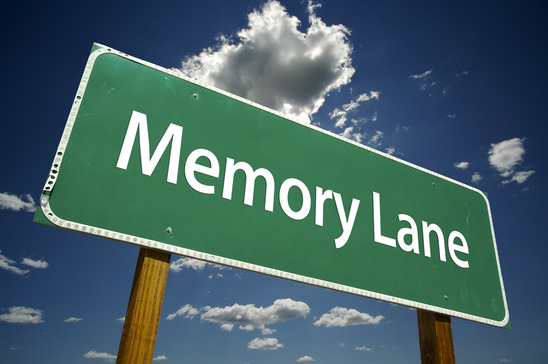 This is an image of a green sign with a white border in the shape of a horizontal rectangle. The sign reads ‘Memory Lane’ in white letters. The background is blue sky with white clouds scattered throughout.