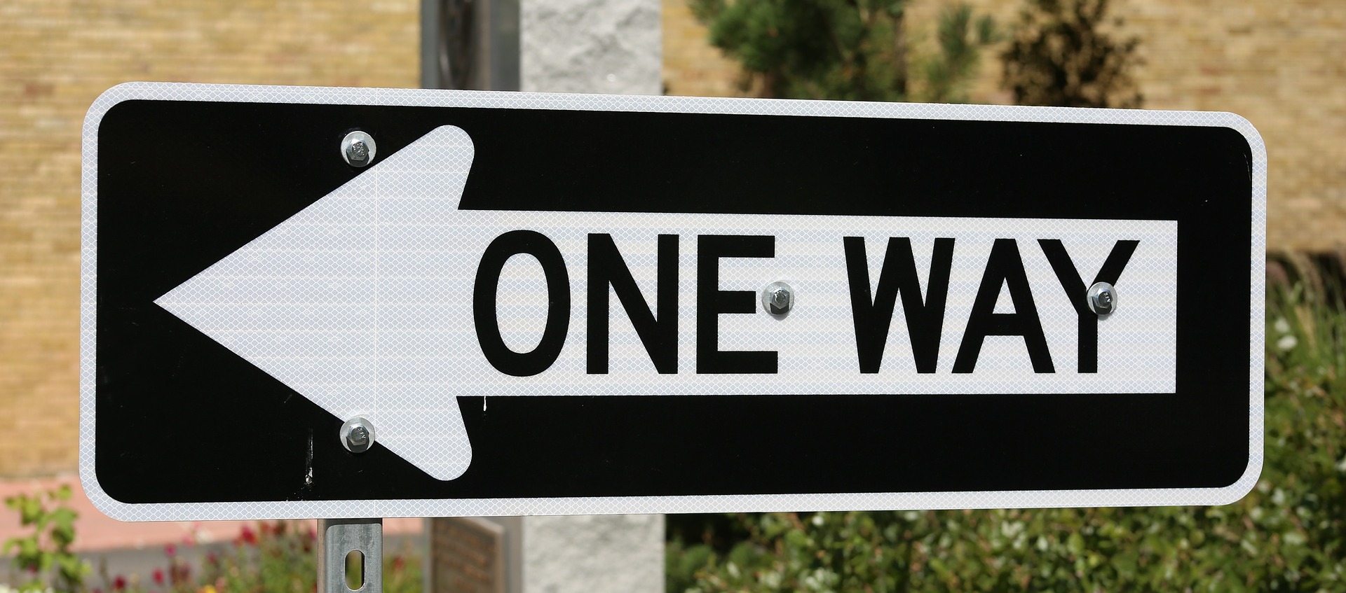 This is an image of a rectangular sign in a horizontal position. The sign is black with a white border. A brick wall and small, green bushes are in the background. On the sign is a white arrow pointing to the left with the words ‘one way’ written in black letters.