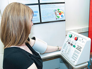 This is an image of a woman using a blood pressure monitoring machine. The woman is visible from the shoulder up, and she is at the left of the image. She is wearing a blue, short-sleeved shirt and has blond hair. Her arm is through the inflatable cuff portion of the machine. The machine is in front of her on the right side of the image. The face of the white control panel has instructions written on it, a green button at the top, with a red button to the left of the green button. The background is a white wall with informational posters on it.