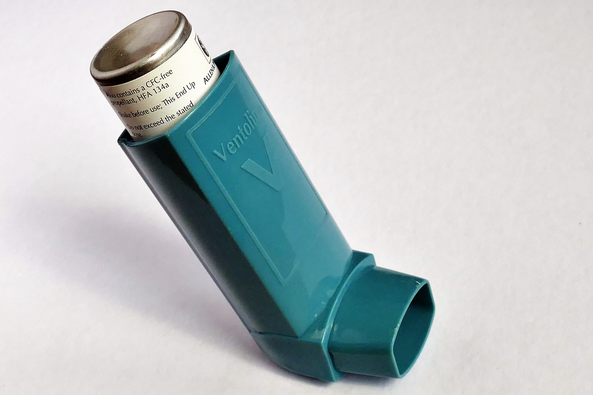 This is an image of a blue asthma inhaler on a white background. A raised ‘V’ with the word ‘Ventolin’ written above it is on the front of the inhaler. A metal canister is visible sticking out the top of the inhaler with a white label on it.