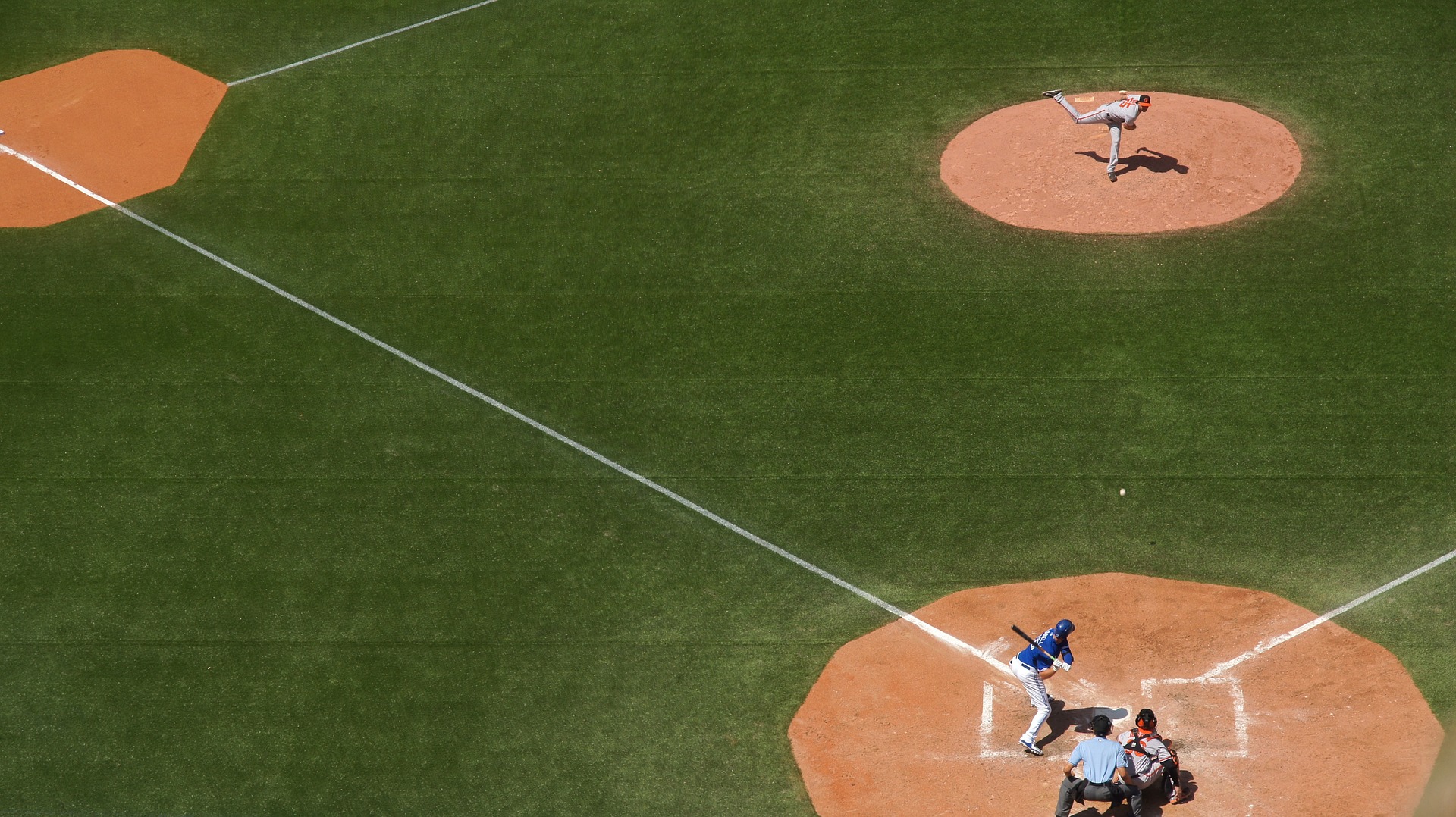 This is an aerial image of a grass-covered baseball diamond. The home plate is in the lower-right corner, third plate is in the upper-left corner, and the pitcher’s mound is at the top-right. The baselines between third and home is clearly visible as a straight, white line. The pitcher is in a white uniform and has just thrown the baseball. The batter is in position to swing at the ball, and is wearing a blue shirt and white pants. A catcher wearing a white uniform is crouched behind home plate, and an umpire in grey is crouching behind the catcher.
