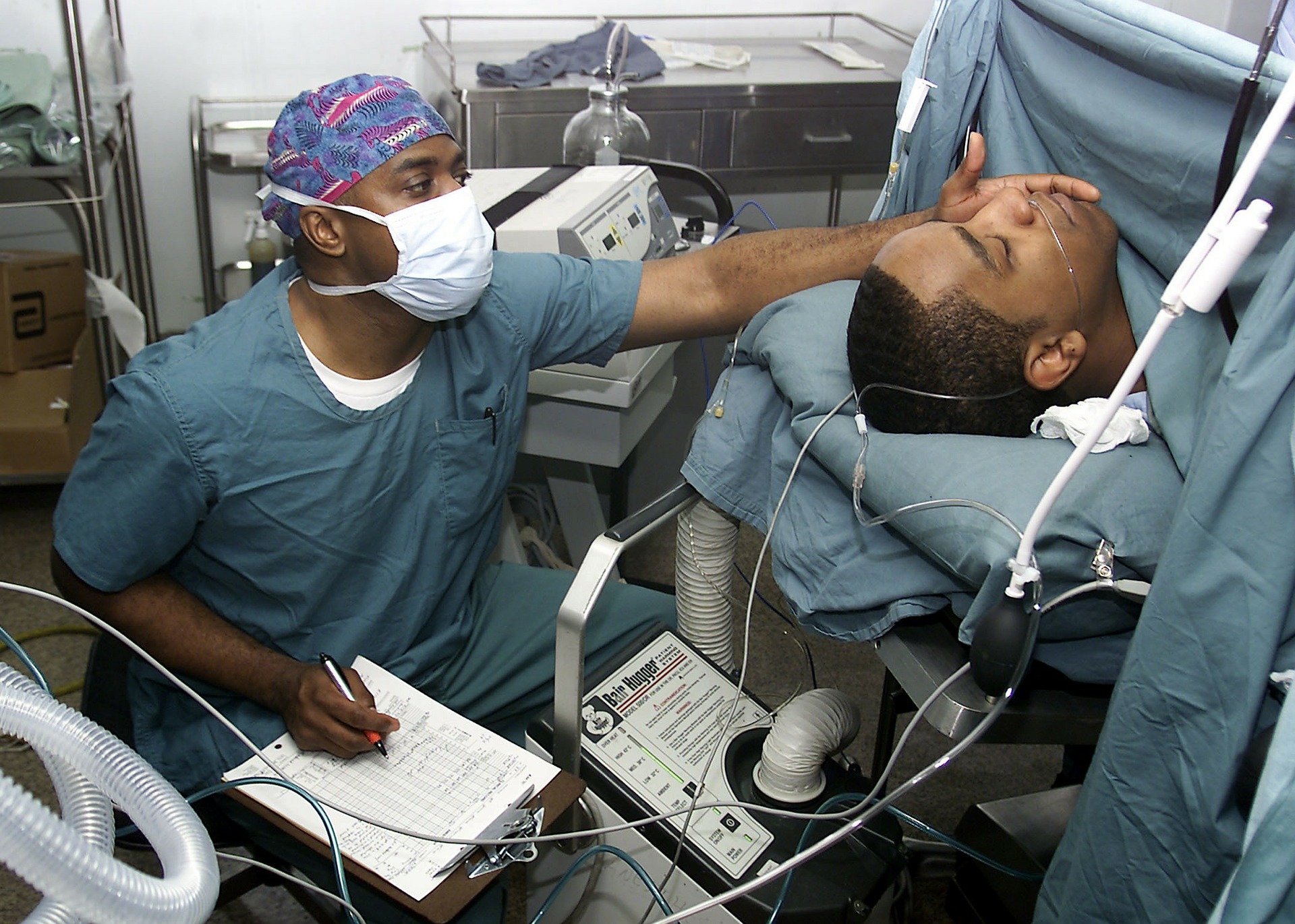 This is an image of a medical professional in blue surgical scrubs sitting on a chair in an operating room. He is wearing a blue and red cap and a white surgical mask. On his lap is a clipboard with white paper on it. He is surrounded by various medical machines. To his right is a operating table with a man lying down on it. Just his head is visible, as there is a surgical curtain across his neck. The patient has oxygen tubes in his nose and his eyes are closed. The medical professional is reaching up to the man’s face with his left hand.