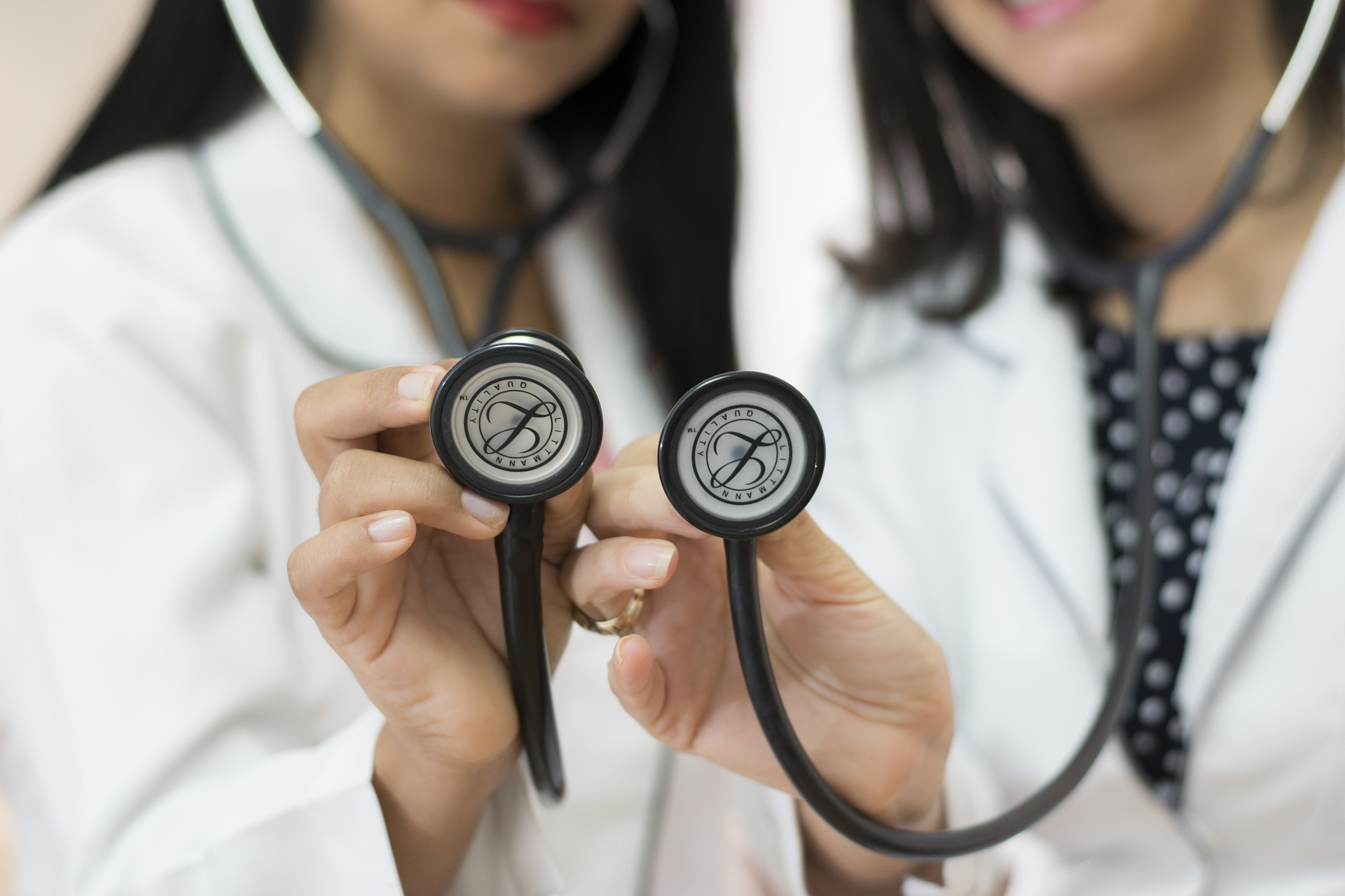 This is an image of two doctors, each wearing a black stethoscope.   Both are wearing white lab coats and have long black hair. The woman on the right is wearing a black, polka-dotted shirt under her jacket. The image shows the women from the waist to the mouth. The women are in the background and are blurred, but each is holding up the bell of the stethoscope in the centre of the image and those are in focus.