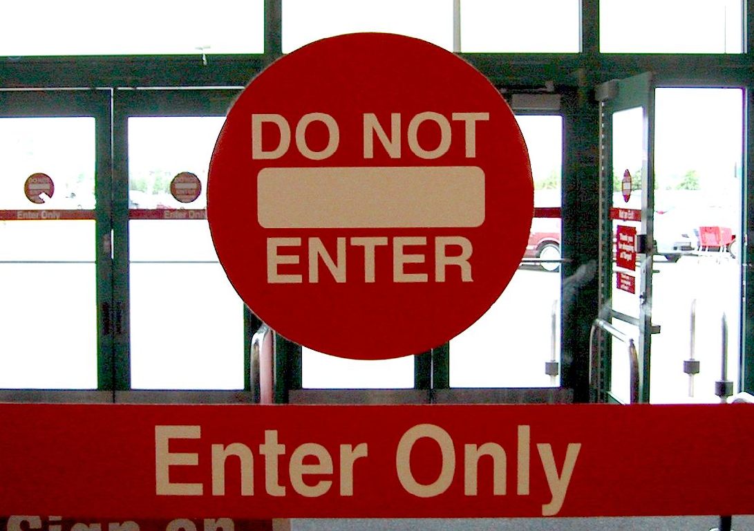 This is an image of two signs on a glass entrance to a store. Doors to the parking lot are visible in the background. The top sign is round with a red background. It reads ‘DO NOT ENTER’ in white letters, and has a white horizontal bar in the centre. The sign below is a horizontal rectangle also with a red background. This bottom sign says ‘Enter Only.'