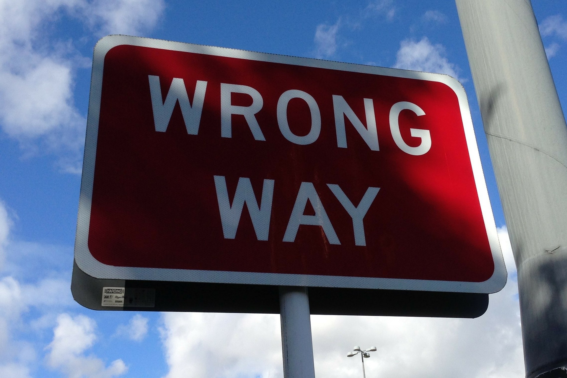 This is an image of a rectangular sign with a red background and a white border. The sign says ‘WRONG WAY’ in white letters. The background is blue sky with wispy white clouds scattered throughout. A grey pole is visible to the right of the image.