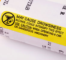 A close up of the side of an orange medication vial from a pharmacist. Overtop of the prescription label is a yellow warning sticker that reads: May cause drowsiness. Alcohol could intensify this effect. Use caution when operating a car or dangerous machinery.