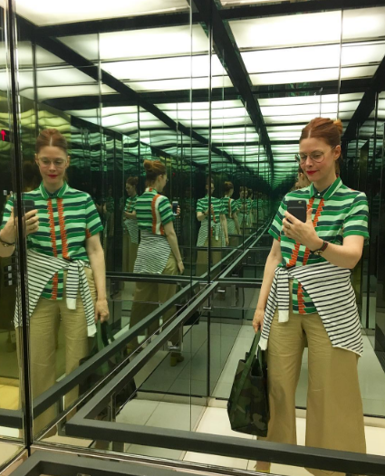 This is an image of a young woman wearing a green striped shirt and khaki pants. She is standing in a mirrored elevator and taking a photo of her own reflection, which in turn is being reflected again an infinite number of times.