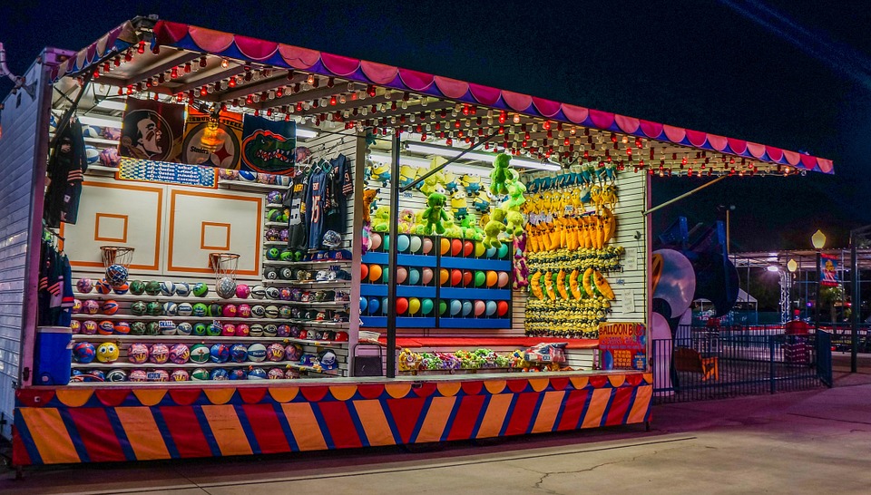 This is an image of two games side by side in a carnival midway. The game on the left involves shooting a basketball into a net. The game on the right involves popping inflated balloons with a dart.