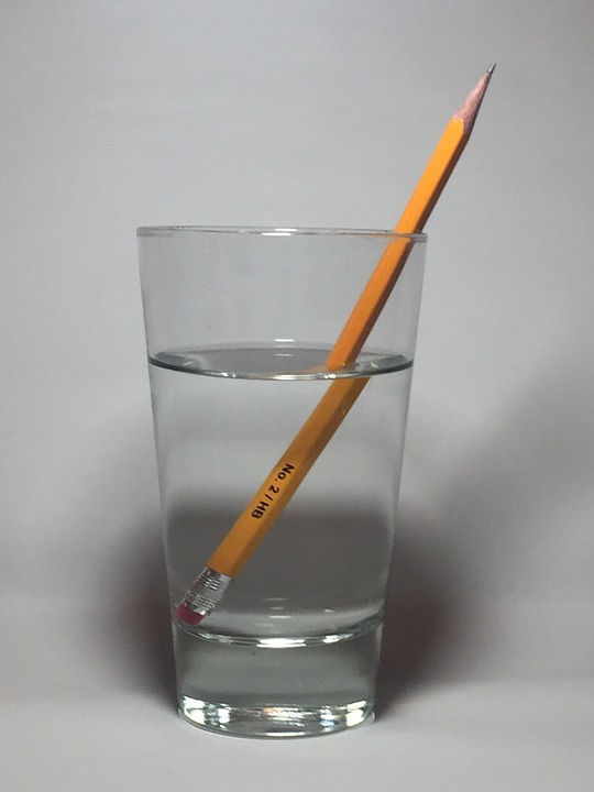 This is an image of a clear, colourless glass of water containing a pencil. The pencil appears larger in the water and to be disconnected from itself at the air and water interface. The glass is also magnifying the pencil to a lesser degree.