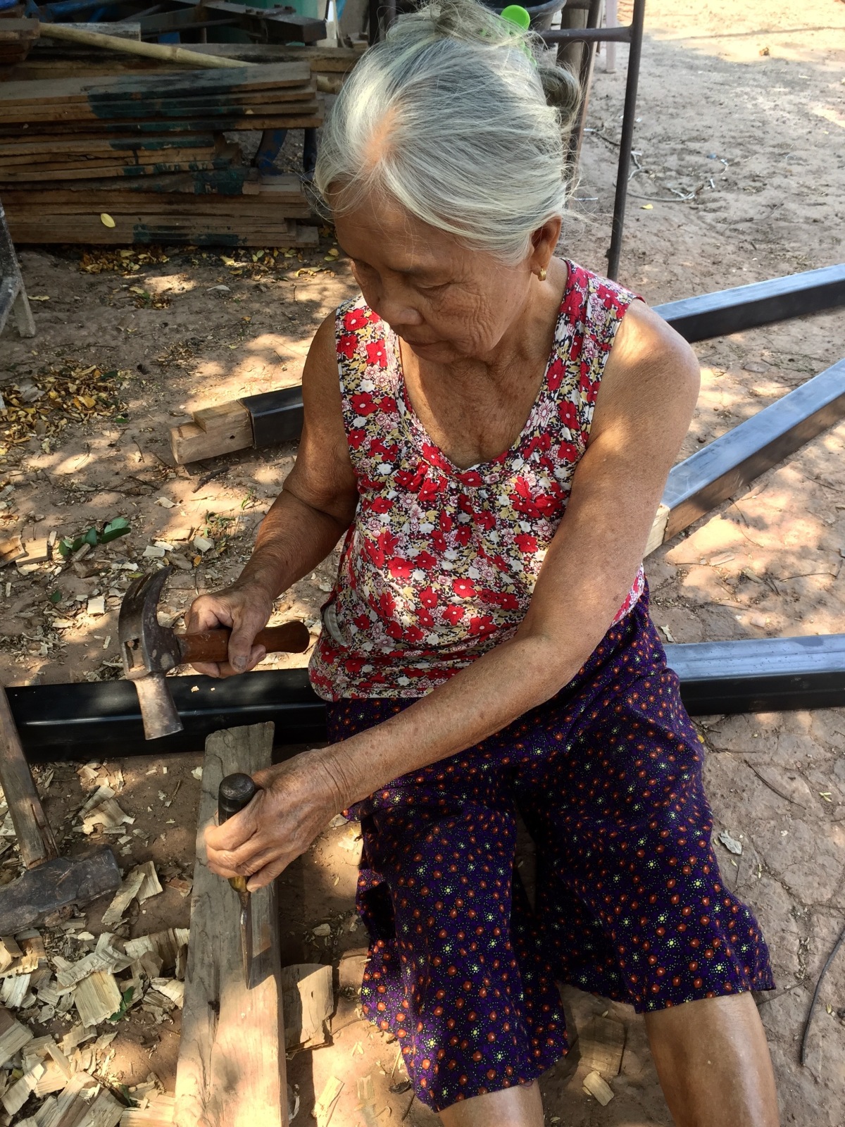 This image depicts an elderly Asian woman, wearing a flowered red and white tank top and blue shorts, using a chisel and hammer to cut a piece of wood.