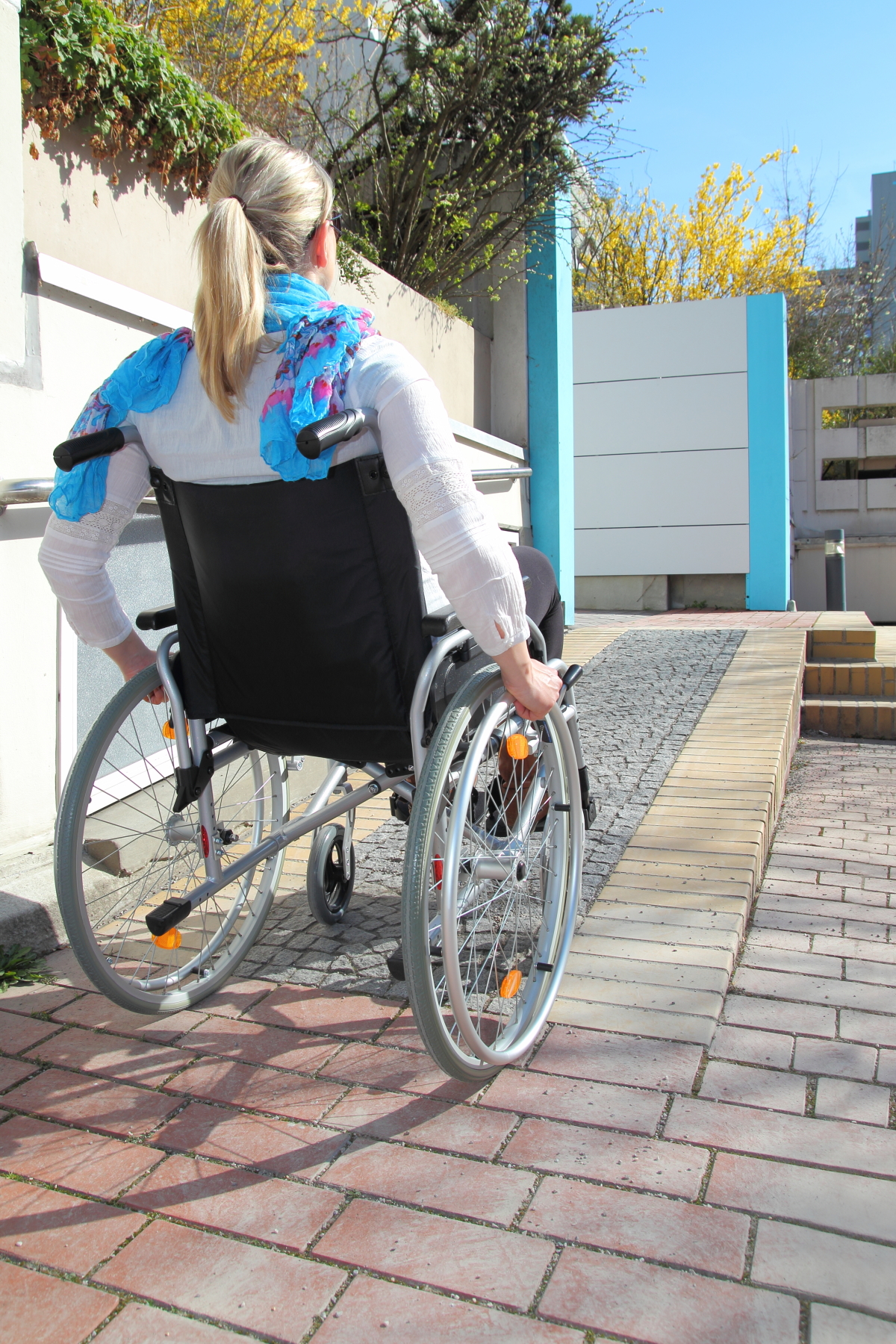 This image depicts a woman with blonde hair in a ponytail, wearing a blue scarf with a white long-sleeved shirt, moving up a ramp in a wheelchair on a red bricked ramp.