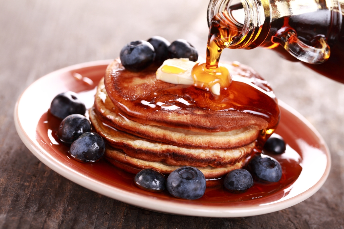 This image depicts maple syrup being poured onto a stack of pancakes and blueberries. 